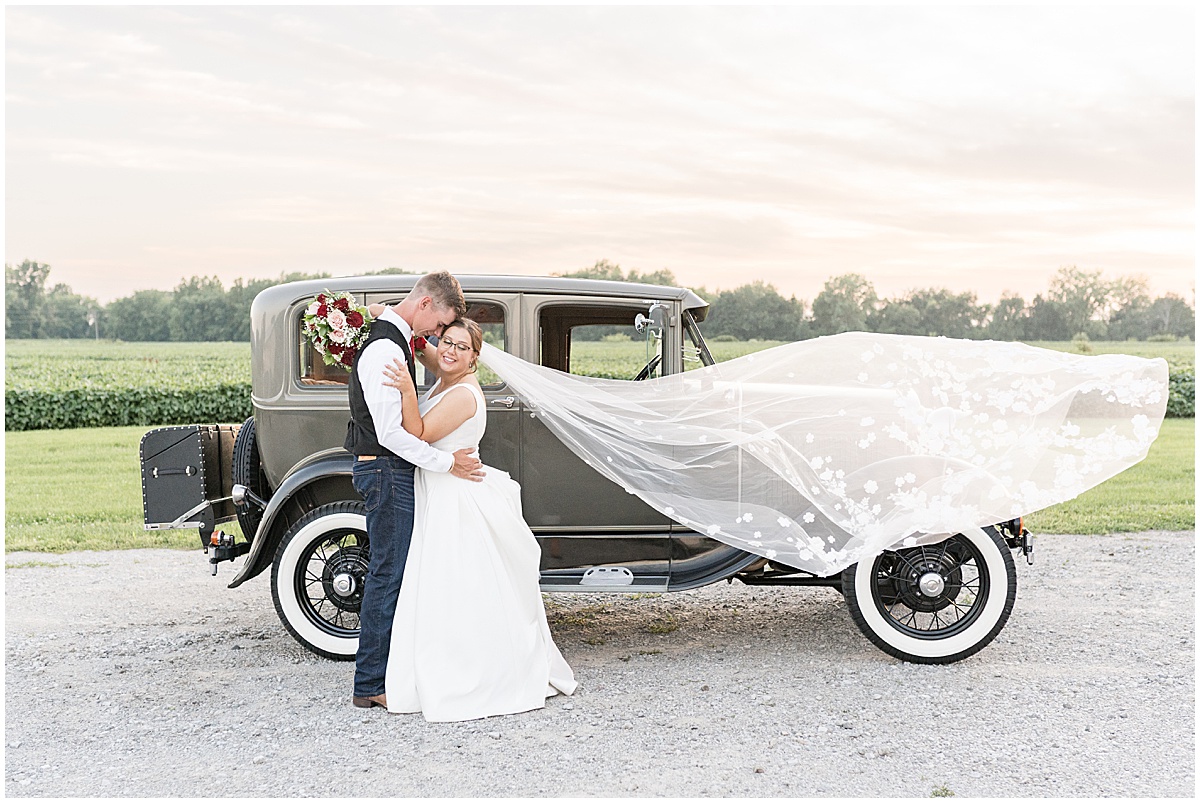 Bride's veil blows in wind in front of vintage car at Churchill Farms wedding in Lake Village, Indiana